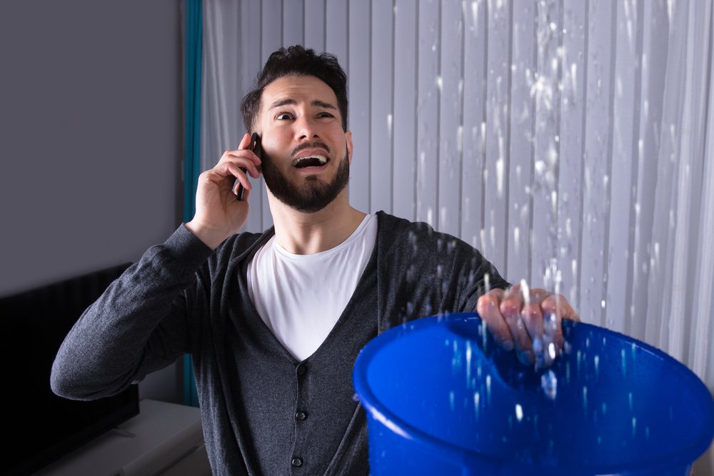 Worried Young Man Calling Plumber While Leaking Water Falling Into Bucket